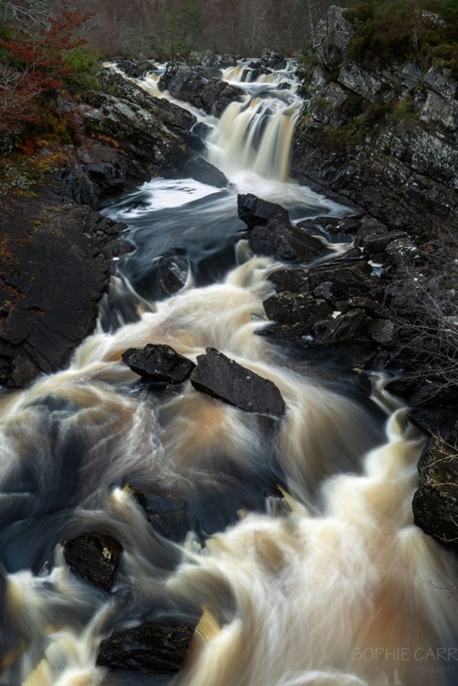 A waterfall with tannin-rich water falling over black rocks. Some autumnal trees just visible, as well as a really annoying white frothy bit at the foot of the main falls.