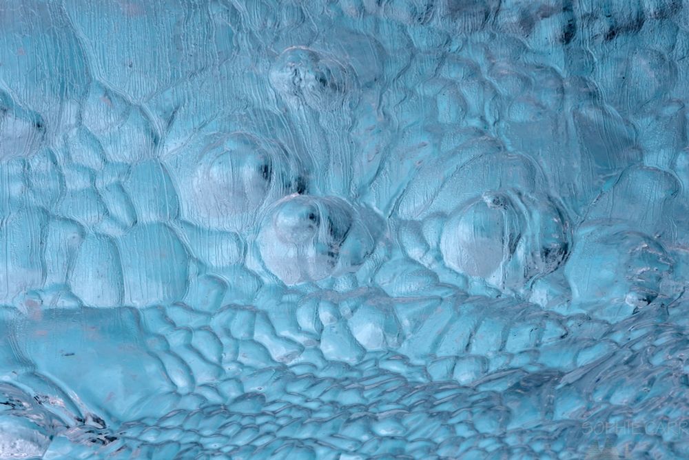 Close up abstract of the surface of an iceberg. It is turquoise with some pink light, and the pattern looks like bulging convex scallops, but is actually convex dips.