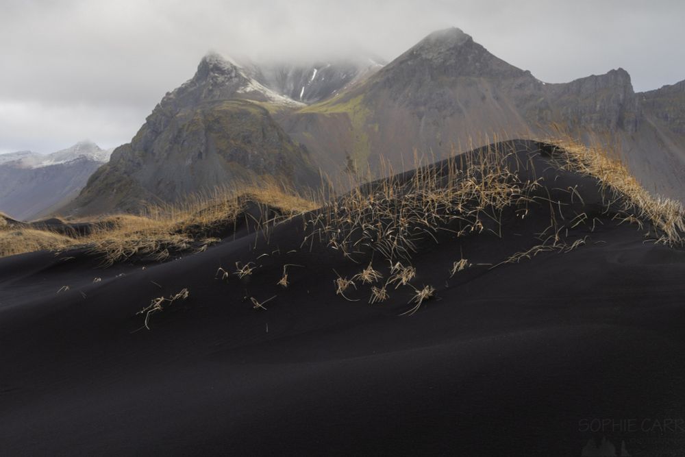 A black sand dune with some yellow grasses sits in front of some mountain peaks, which have some cloud cover at the top and a touch of snow on the top left peak.