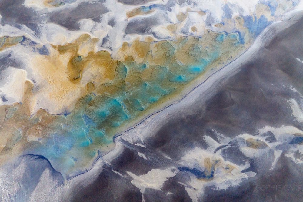 A weird image with the bottom right hand side grey sand with some pale patterns in it. On the top left side there are some orange bits of sand (as well as pale and grey) and in the middle are some turquoise pools.