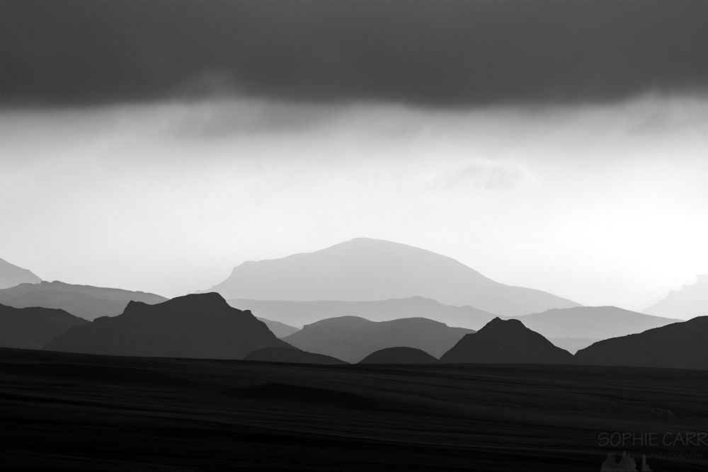A black and white image with lots of layers - at the top dark cloud, then a lighter area of sky which has some vaguely visible clouds, then layers of mountains, hills and sand in the foreground, each getting darker towards the front of the photo. There are two distinctive jagged pointy triangular mountains but the rest are generally rounded and more flat.