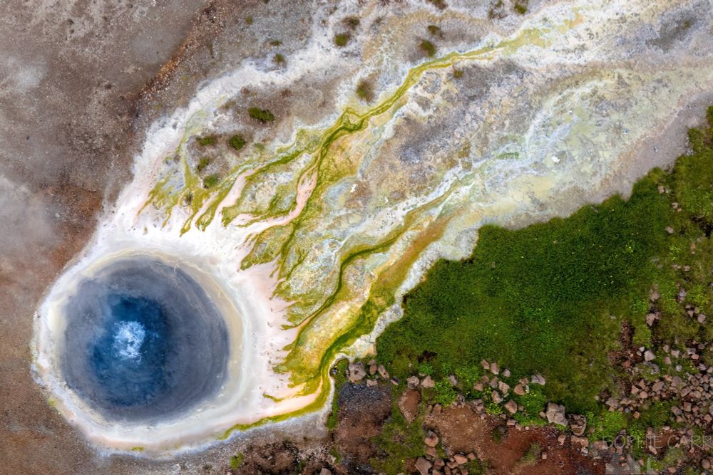 A geothermal hot pool photographed from above. The pool is in the bottom left of the photo and is blue with white surrounds and a bubble in the middle. From it - stretching out to the top right - are streams that are pale orange  and then green and white patterns leading outwards. There is some grass to the bottom right and pinkish stones, and the ground top right is pinkish-brown.