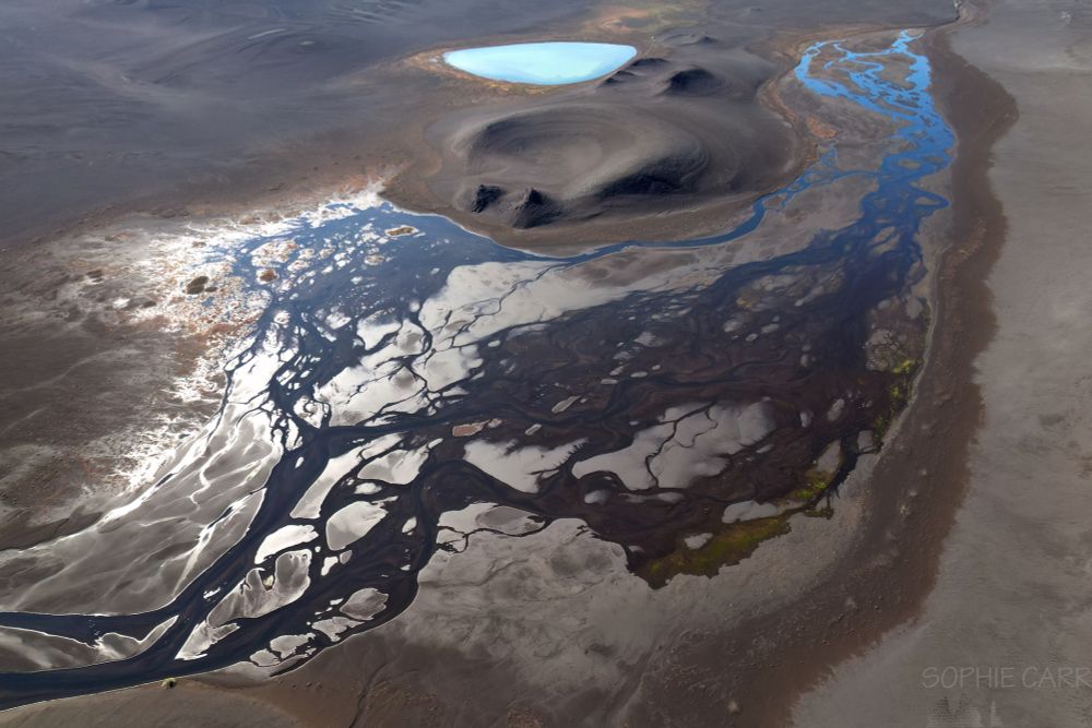 An aerial view of a braided river meandering through the Highlands of Iceland, with the landscape predominatly brown sand. In the distance are a couple of low eroded volcanic craters and a turquoise lake at the top. The river is generally dark, but blue at the top where the light shines on it.