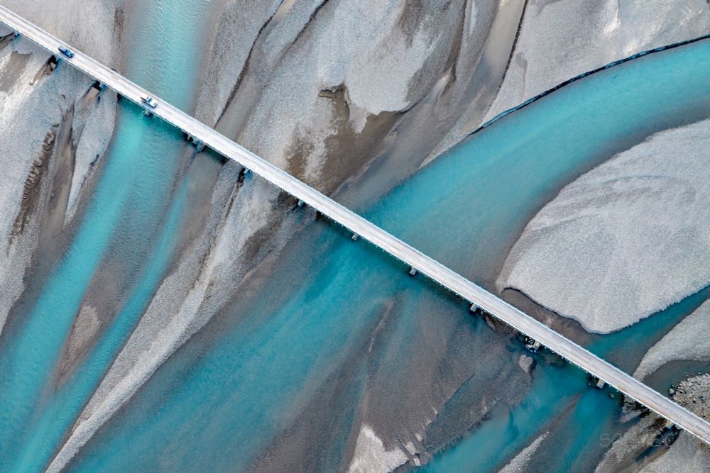 A grey road bridge crosses a river plain with grey sand and bright turquoise river braids. There are a couple of cars at the top of the bridge.