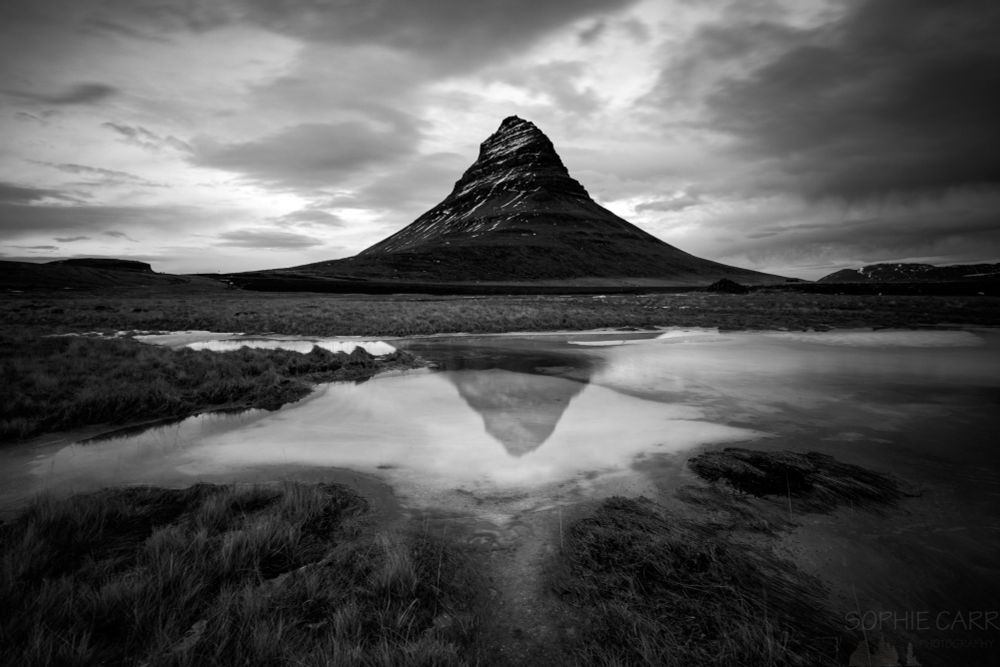 A black and white photo of a very pointy mountain in the middle of the shot, reflected in a frozen pool below. There is some cloud around and some grasses in front of the pond.