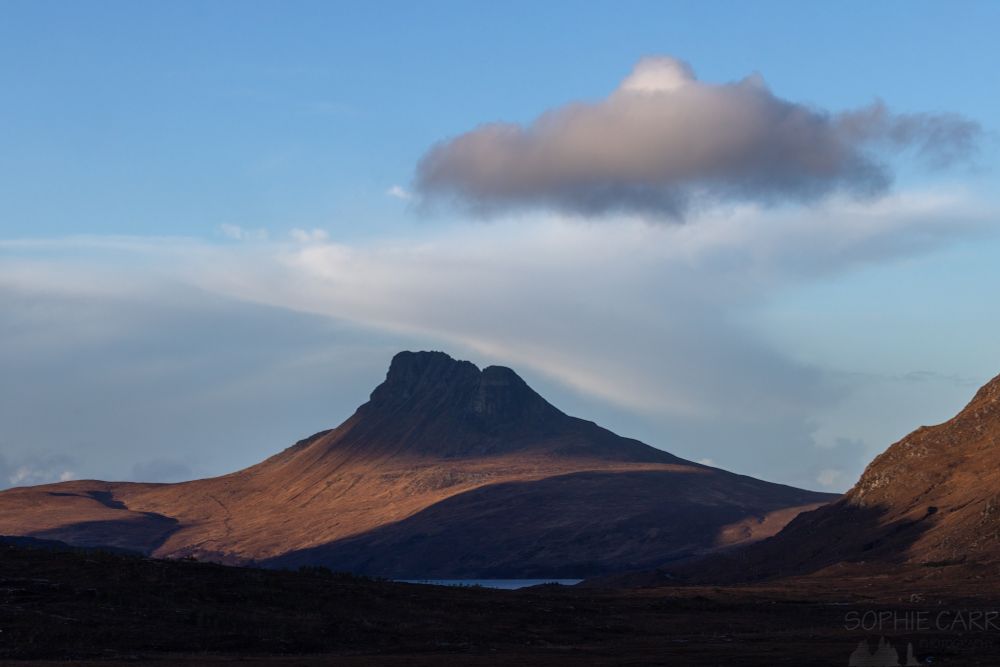 A pointy craggy mountain in the distance with the peak under shadow. Below is a strip of light illuminating the orange vegetation. Above is a hat shaped a bit like a sombrero.