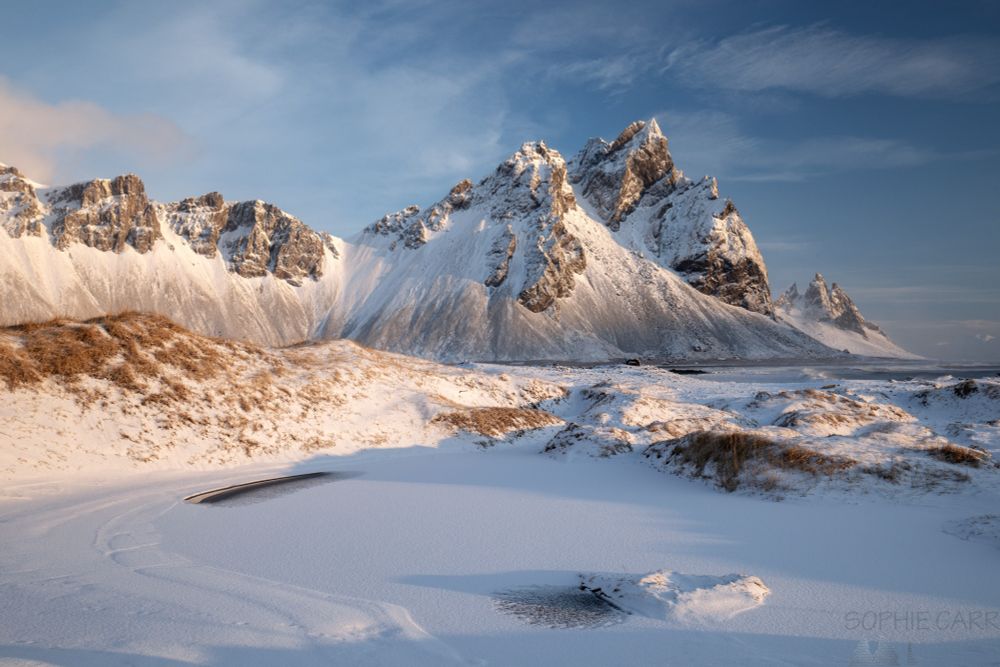 A snowy scene in low sunlight with a snow-covered pond in the foreground, some snow-covered dunes in the middle, and snow-covered pointy rocky peaks in the distance. Blue skies with some wispy clouds above.