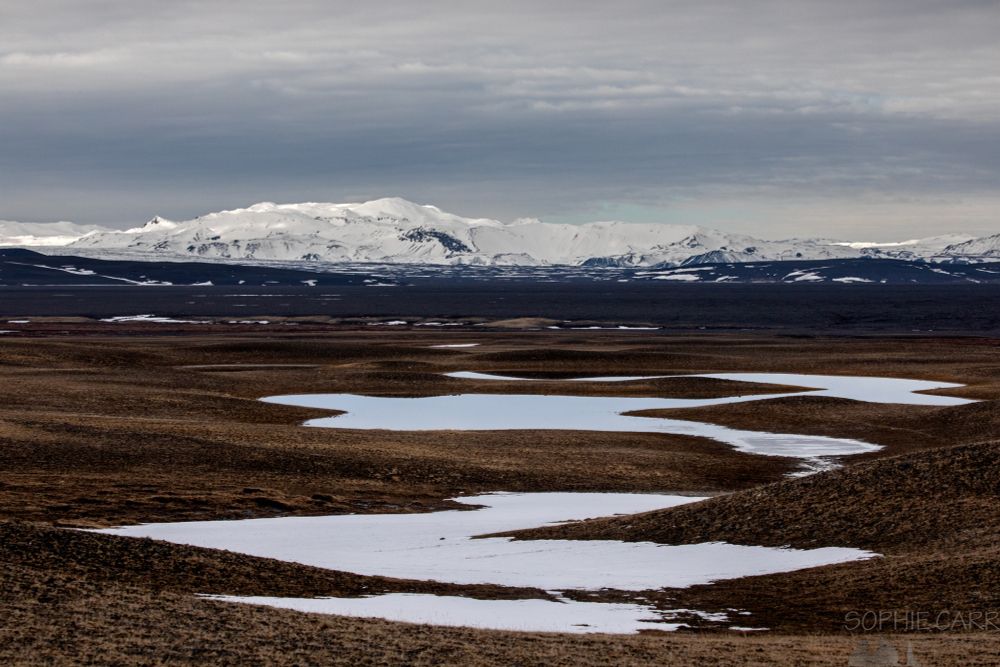 A view of brown landscape with some icy ponds in interesting shapes disappearing into the distance. There are some snowy peaks on the horizon, some with some sunshine on them. A band of grey and white cloud hangs above. Looks autumnal but it's taken in late winter.