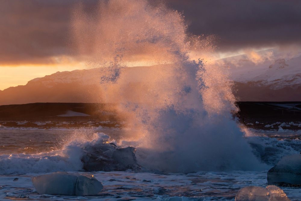The tail of a crashing wave shoots into the sky after breaking over icebergs in the surf. The spray is illuminated by the setting sun (out of sight off to the left of the shot) but glowing pink. Behind are some low mountains and some dark cloud above.