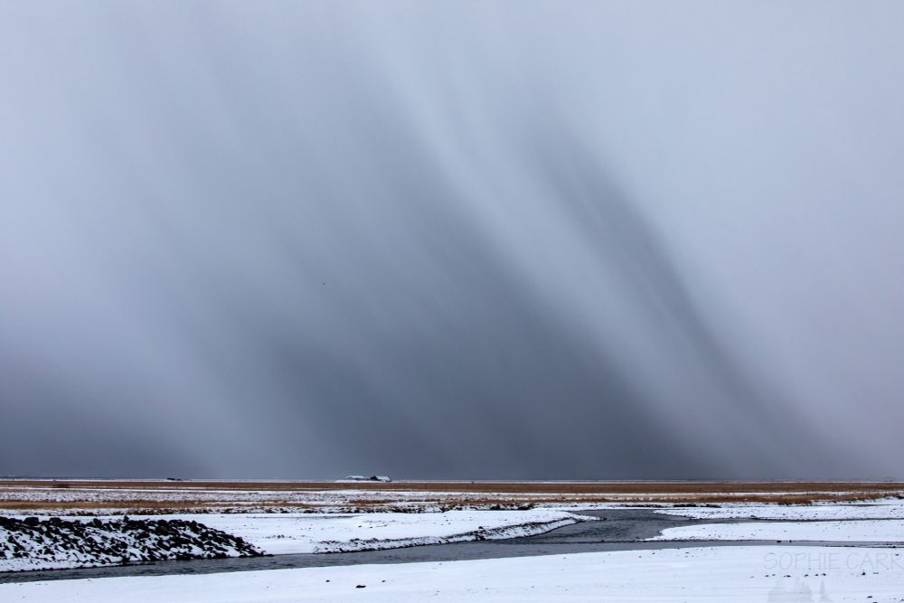 A massive snow storm visible in the distance. In the foreground in the bottom 1/4 of the shot is a wintry scene with a grey river, snowy banks, a couple of houses on the horizon. Above are enormous visible snow storm clouds - with stripes of dark grey shooting downwards to the horizon.