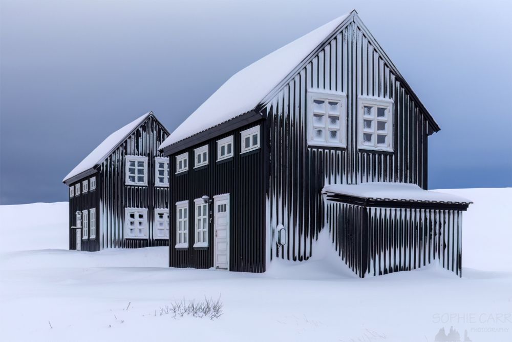 Two Scandinavian-style black-painted wooden houses with white-painted windows and doors covered in snow. There is snow on the ground in the foreground - with a few grasses showing. Above the houses the sky is a dark, ominous stormy blue on the horizon; a little lighter above. There are snow drifts against a little shed attached to the nearest house, and snow has built up in the window panes too.