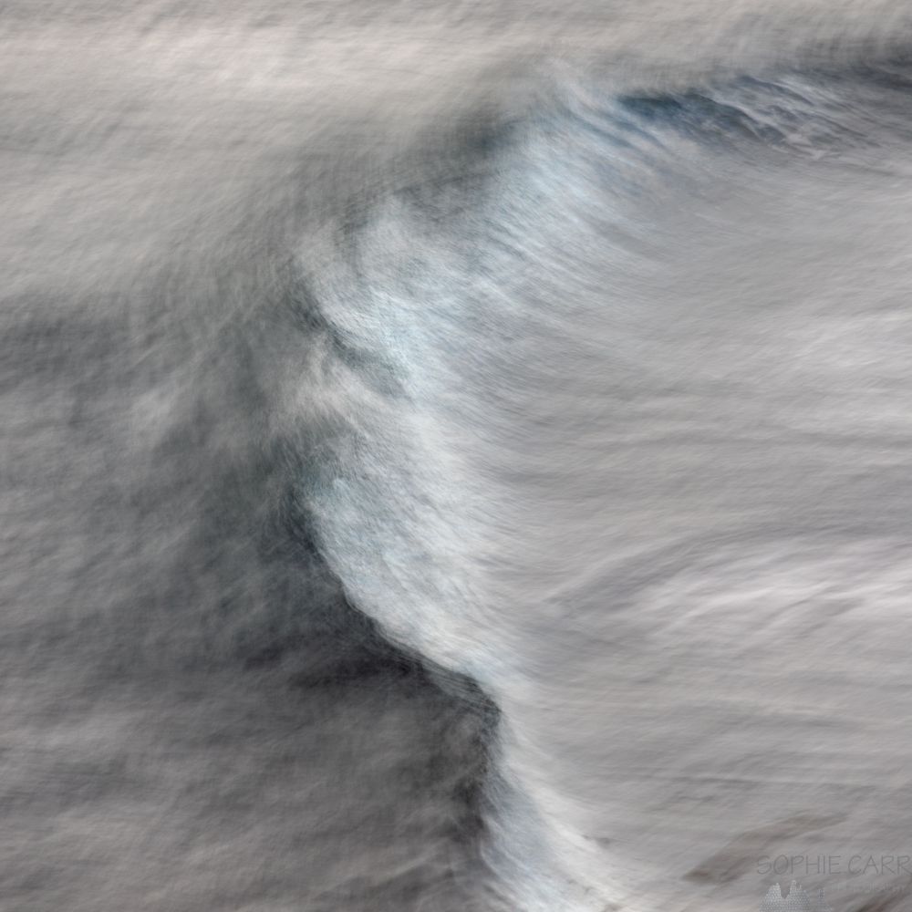 Long exposure, hand-held shot of a wave breaking. Square crop of a photo taken from above. Mostly grey, with a curved wave through the middle, with darker grey, pale turquoise and white touches.
