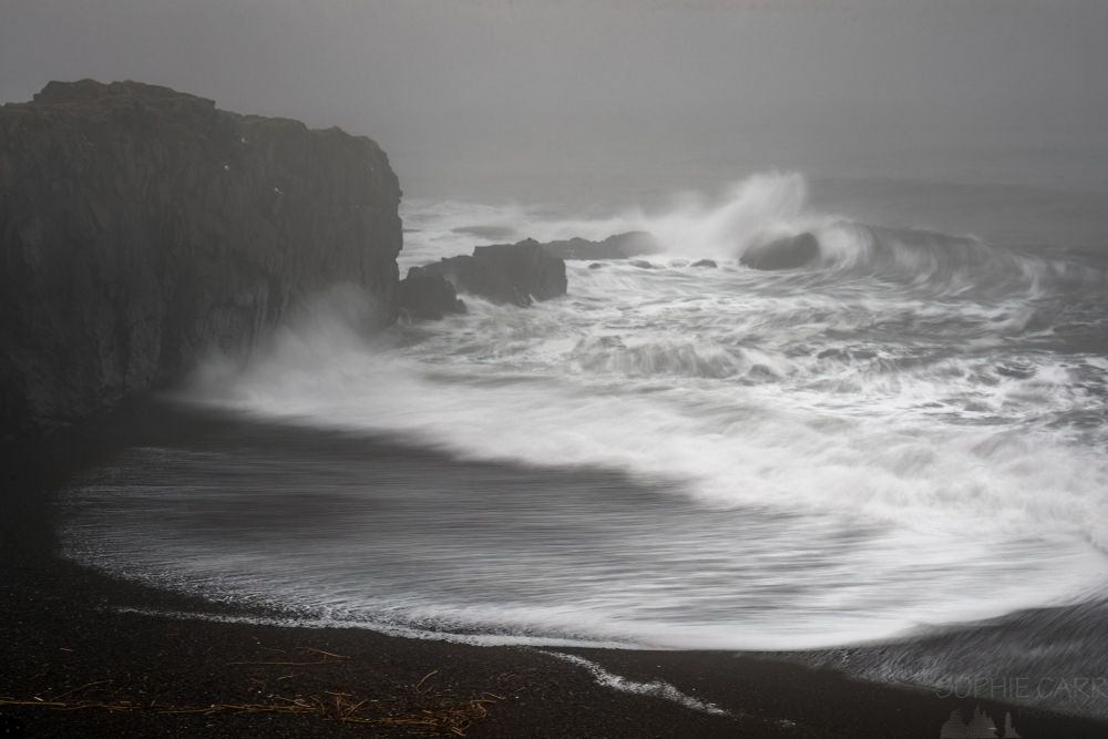A foggy scene with a black sand beach with a black rocky cliff jutting out into the sea. There are white waves crashing against rocks and the beach. The exposure length is slowed down a little to give some smoothness to the water movement.
