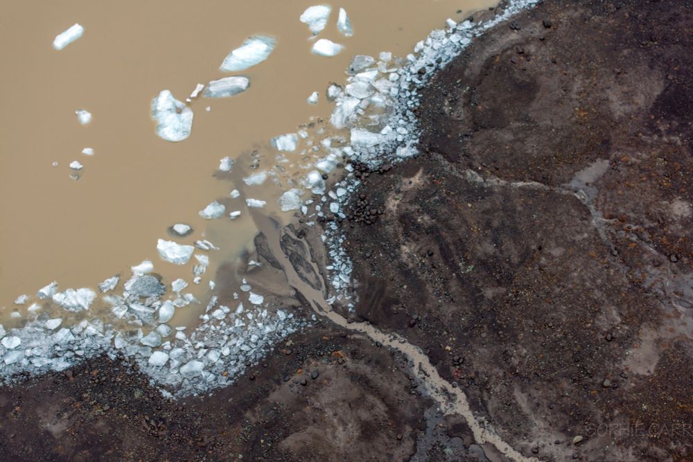 A drone view looking down to the edge of a lake full of milky tea brown water with tiny bits of ice dotted along the shore. The land is brown with a brown river flowing down into the lagoon.