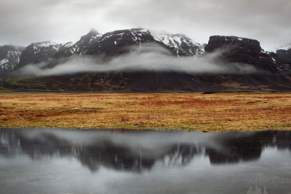A photo of some mountains with a little snow on them reflected in a pond at the side of the road. In the middle is some brown and red grasses (as it's late winter) and there's some puffy fog patches over the mountains, also reflected. Grey and white skies above; miserable weather.