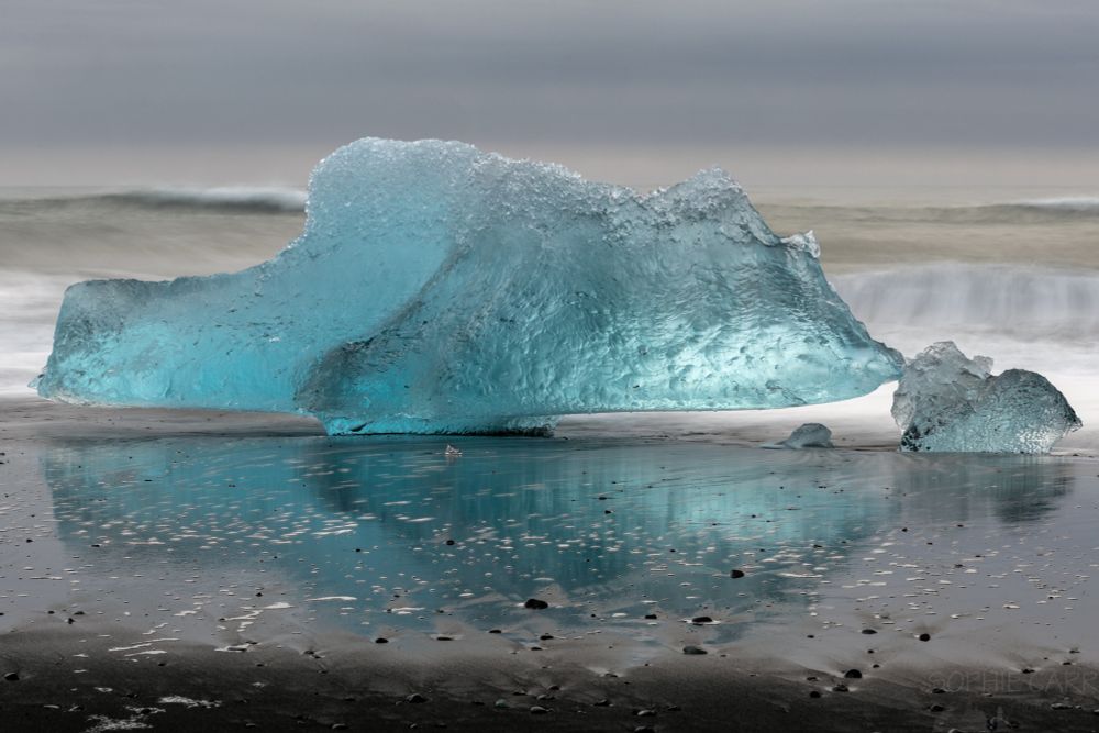 A very bright turquoise iceberg, with a couple of little pieces to the right, sits on a black sand beach and is reflected in the wet sand. There are some long exposure smooth waves behind and layers of grey sky above.
