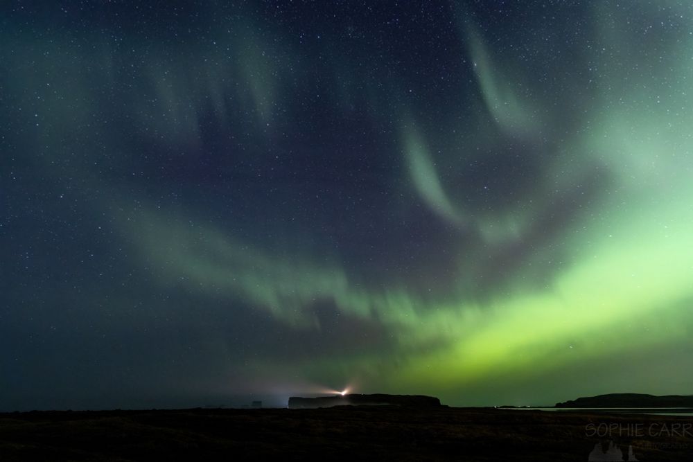 Green northern lights across a starry dark blue sky. There is a smudgy green area on the right with some streaks to the left of the photo. Below is the silhouette of a promontory with a lighthouse on the top. A little lake can just be seen with a green hue at the bottom right.
