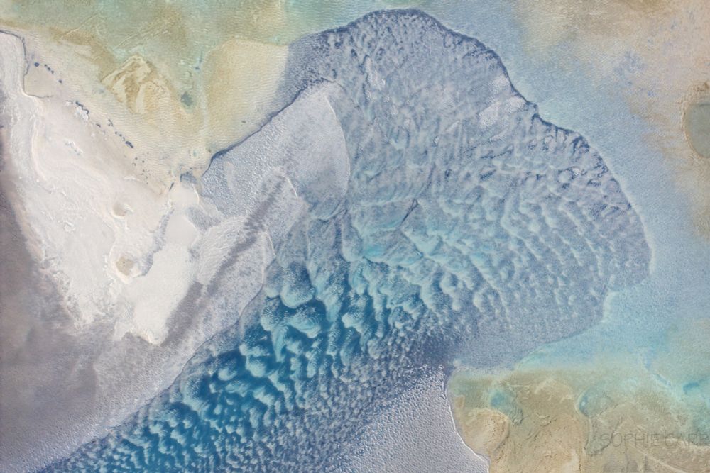Drone view of some weird patterns in a lagoon near the coast. There are some turquoise patterns that look very similar to the scalloped patterns of an iceberg. There is some yellow white, pale brown surface around the central turquoise shape.