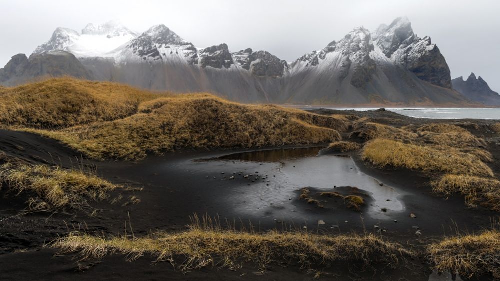 An almost dry pond sits in black sand dunes with some yellow grasses on the dunes. In the distance is a huge mountain range with jagged peaks and a light covering of snow. A very pointy mountain - Batman-shaped - is just visible on the right.