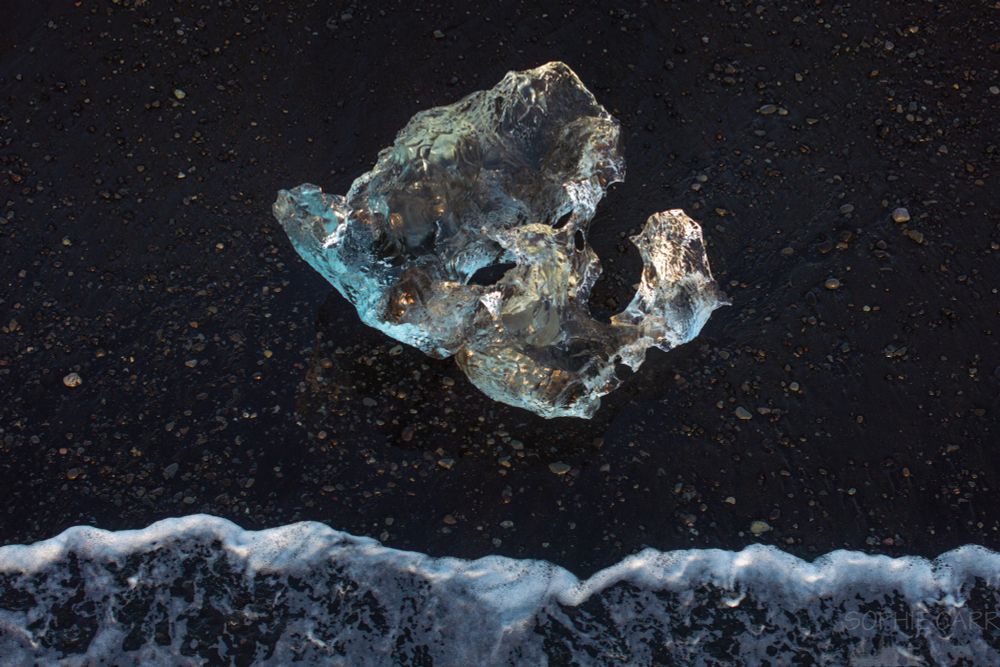 A lone iceberg on a beach photographed from a drone above. At the bottom a wave is coming in towards it up the black sand and pebbly beach. The iceberg is mostly turquoise but there are some yellow patches from the reflection of the sun low in the sky. It looks a bit like a smiling face mask.
