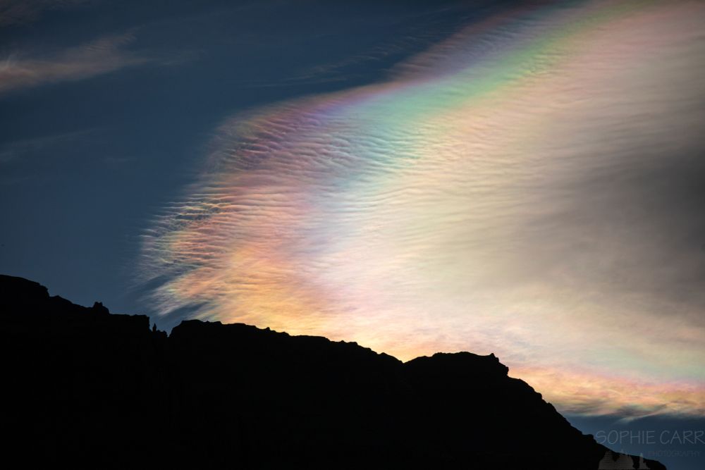 A dark jagged silhouette between some very colourful (rainbow) nacreous clouds.