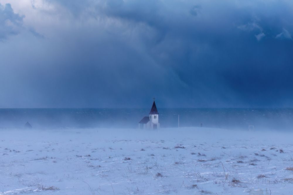A little church is sitting in a snowy field with blowing snow going past and huge blue clouds above. The church is boarded up and lonely-looking, with a dark pointy spire and roof (which are red, but you can't see that in this light).