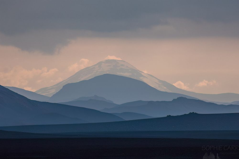 A moody image with about 13 layers of landscape leading off into the distance. The furthest away one is a partially-snow-covered mountain with a rounded top. The other layers are quite blue and get darker at the front, which is almost black. Above the sky is a little pink with grey layers above and there are some dotted white clouds on the horizon above the mountainsides. This view of layers of the landscape is known as landskein :)