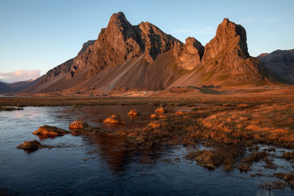 An autumnal scene with some rocky and scree-sloped mountains, and a frozen pond in the foreground with some tufts of grasses glowing orange in the setting sun.