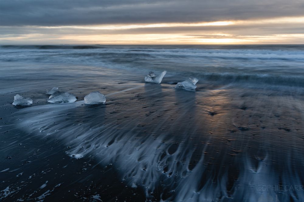 A few tiny pieces of ice sit on a black sand beach. There are some water trails over little pebbles where the wave is receding. There is some sunrise light which is reflecting on the black sand.