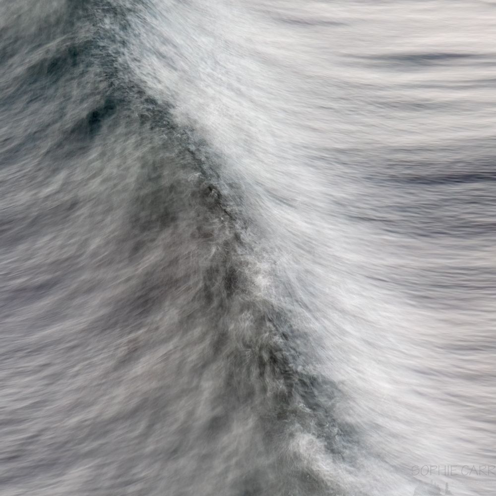 A square crop of a hand-held long exposure photo of a wave taken from above. The photo i s mostly grey, with some dark turquoise and dark grey along the line where the wave is breaking, and lighter grey to the right.