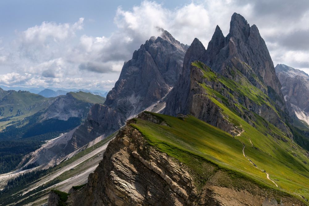 A very pointy ridgeline, with many different peaks jutting up into the air. Below are some grassy meadows in the sun, with a line of people visible hiking across. Below are steep slopes and above some clouds disappearing into the blue sky.