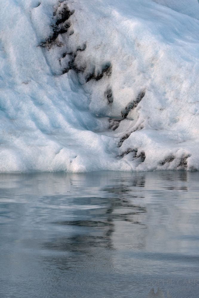 A detail of a mostly white iceberg in muted sunshine, with a touch of turquoise and some black squiggly marks along a seam. This is reflected in the water below and is also squiggly from the vaguely moving water.