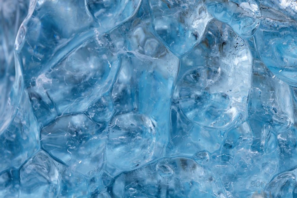 Close up of the surface of an iceberg on the beach. There is a vaguely hexagonal pattern of little convex scallops and the colour is an intense blue/turquoise.