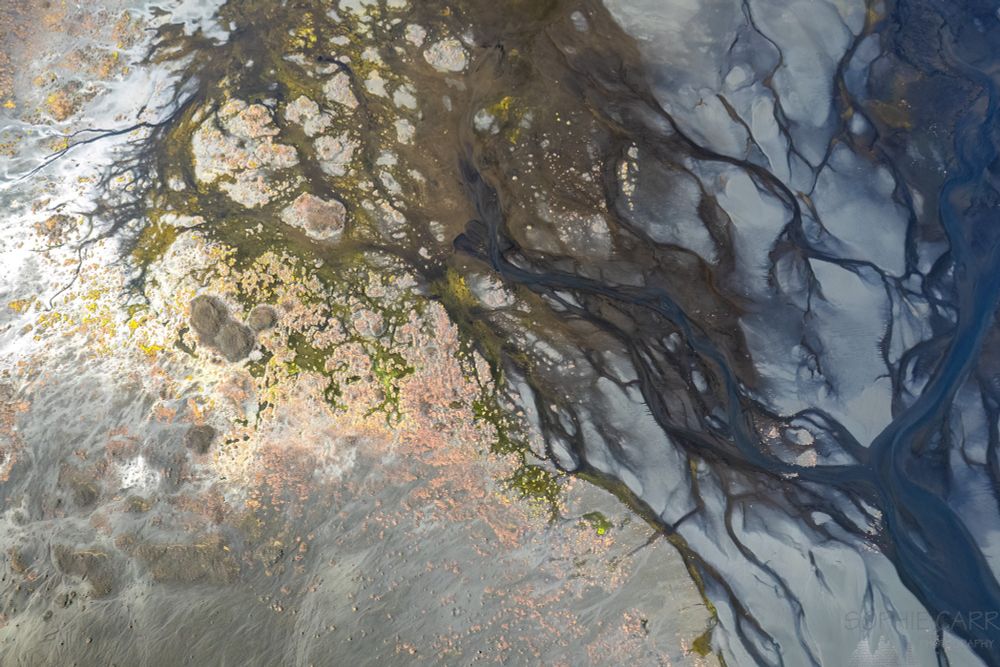 A drone view of a braided dark blue and brown river on the right hand side, with some pale grey sand throughout. There's also some weird red rocks and yellow vegetation on the left hand side.