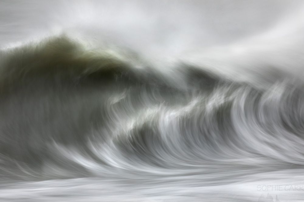 A long exposure of a wave breaking, caught mid-break. The water is greenish, and there are areas of white in the middle of the wave and at the top. Below the water is white and grey streaky, and above is more white spray and some grey ocean behind.