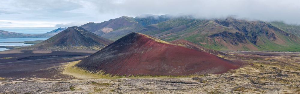 A panorama view of an old very red volcanic crater which is like a squashed pyramid. There's a brown/green one in the distance. In the foreground is lava covered with rather yellow moss and vegetation, and beyond are some grassy mountains with some grey low cloud above. To the left in the distance you can see a fjord and the sea.