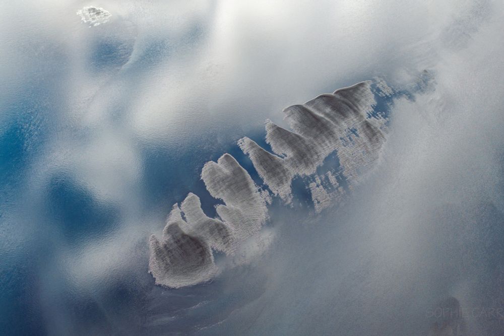 A drone view looking down on an inland lagoon, with clouds reflected in the water's surface. There are some sand banks visible with one large formation that looks a bit like a fish skeleton.