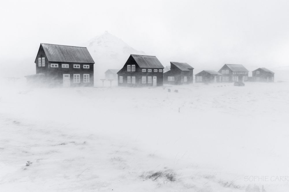 Six houses visible - black painted wooden walls with white doors and white painted windows. The whole scene is blizzardy and monochrome, with blowing snow visible in front of the houses, and some grasses just showing in the foreground. Between the first and second house it's possible to see a pointy mountain.