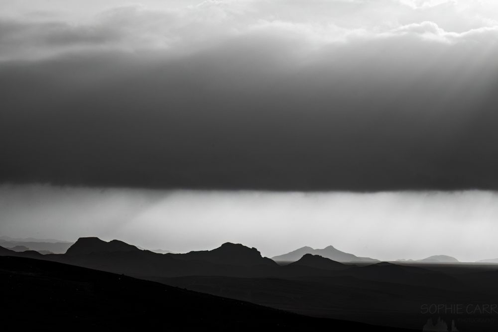 Black and white image showing the silhouettes of layers of hills and mountains at the bottom, a light layer above, then dark cloud band above that and light again at the top. There are some crepuscular rays visible.