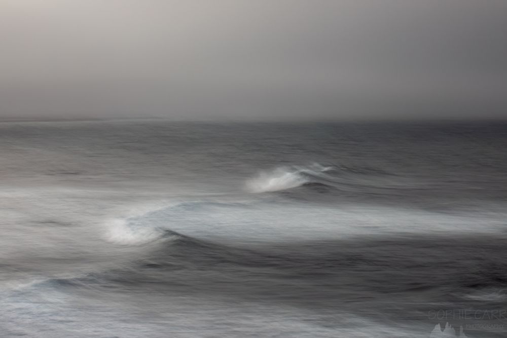 A wider view of waves breaking taken with a longer exposure. The sea is grey, with white froth where the wave is breaking and a touch of dark turquoise. The sky above is grey. 