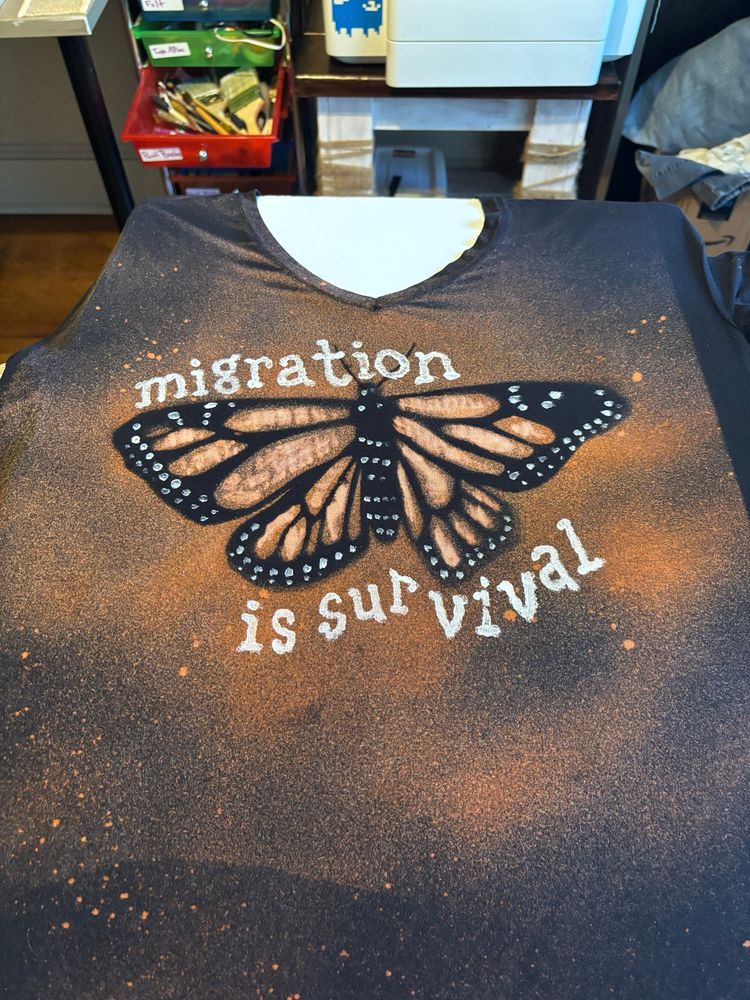 A bleached shirt revealing a monarch butterfly. White letters are painted around: “migration is survival”