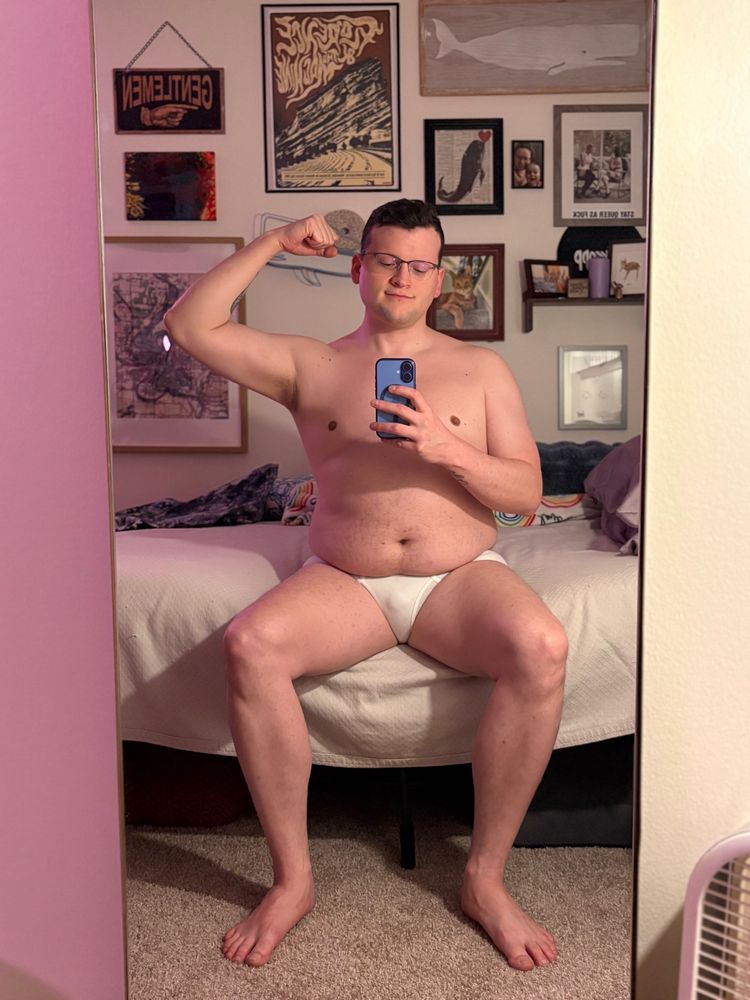 Me flexing my right arm in only white briefs, sitting on my bed facing the mirror