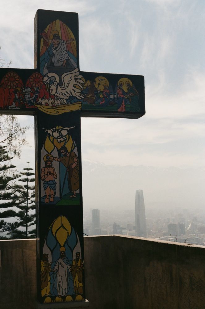 An analog photo of a cross with a painted illustration of several scenes from the story of the Jesus Christ’s crucifixion. In the background is Santiago, Chile. Of particular note is the Gran Torre Santiago—the largest tower in South America. 