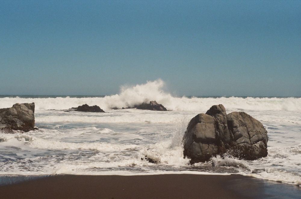 A film photograph of a beach in Chile. Large rock in the foreground, with a wave breaking over another boulder in the middle distance. 