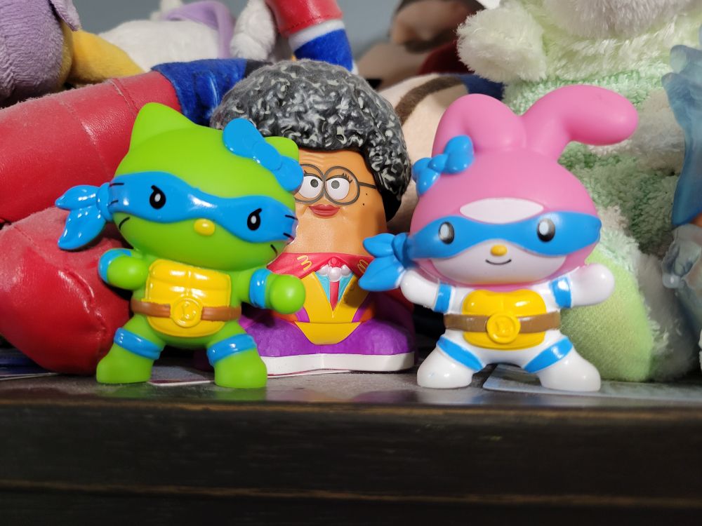 Tiny plastic figures of a green-skinned hello Kitty wearing a blue bandana and blue hair bow who has somehow been morphed with Leonardo the Ninja turtle, and also a my melody with a blue bandana and blue hair bow that has somehow been morphed with the body of Leonardo the Ninja turtle. In the background is a small plastic figurine of a chicken nugget with glasses and a red and purple robe and a lovely afro