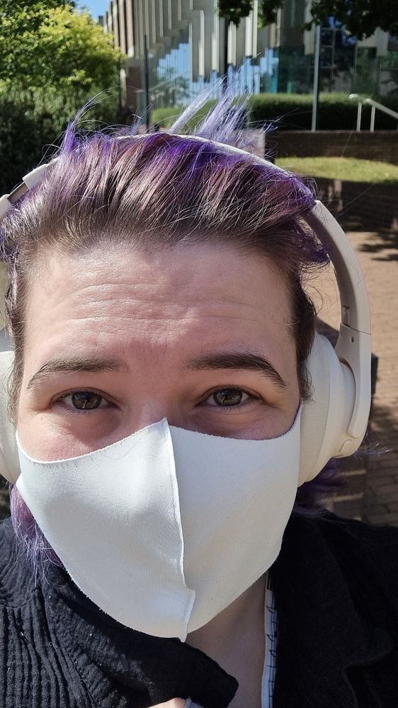 Callie, a late 30s white woman with purple hair, cream headphones, and a white face mask, smiles at the camera in front of a library on a sunny day.