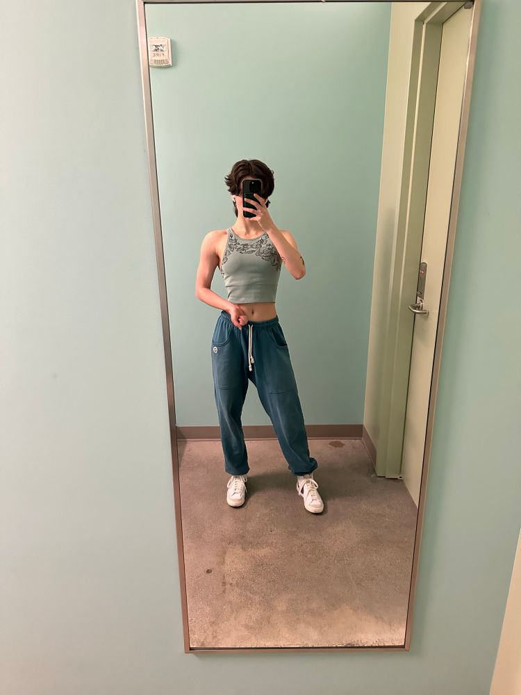 Paul taking a mirror pic after a workout, flexing her arm. She is wearing a turquoise cropped tank top and blue sweatpants