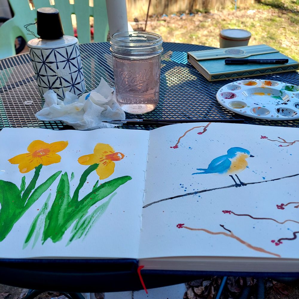 A notebook with two watercolor paintings, one of daffodils, the other of a bluebird on a wire, on a patio table cluttered with painting supplies.