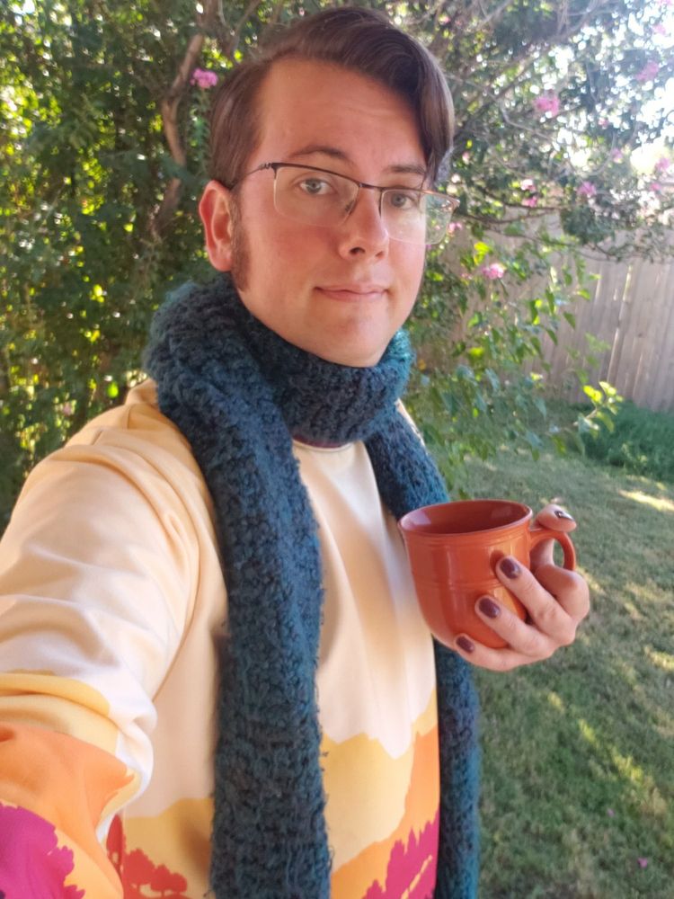 A selfie of me, a cis white male wearing glasses with short brown hair parted to one side. I'm in front of a tree holding an orange mug with nails painted burgundy. I'm wearing a dark blue/green scarf and fall-themed adventure sweater from Paola's Pixels.