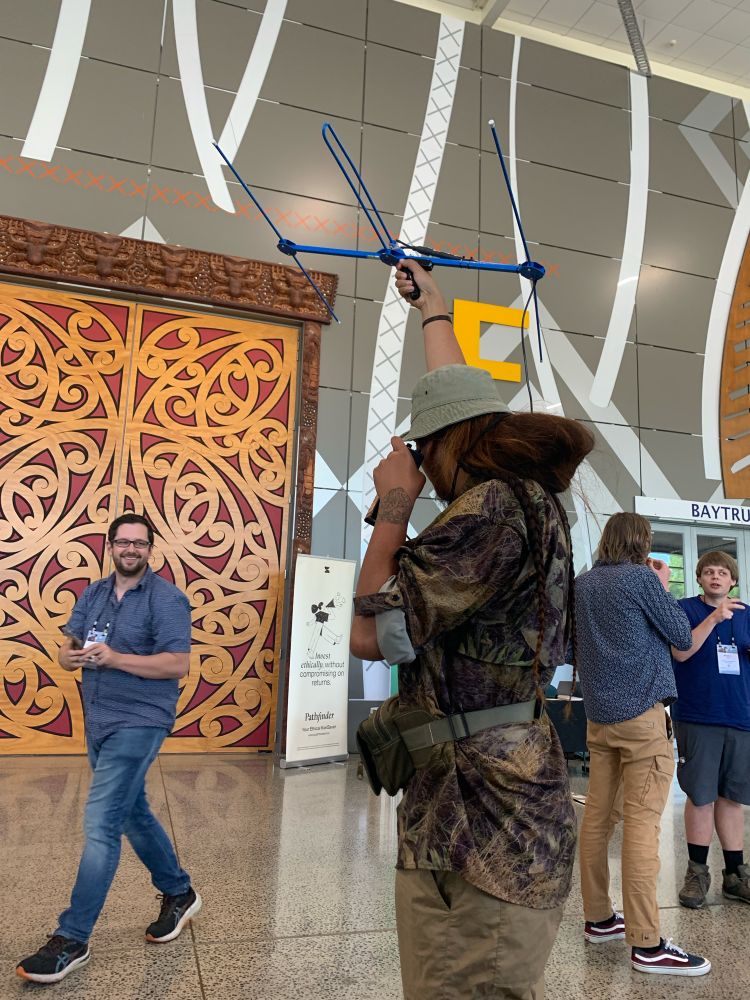 Zion holds up a blue yagi antenna in the air while listening to the receiver, trying to locate the transmitter hidden in the crowd. In the background, in front of the ornate wooden main doors of the meeting room, Ben “Batman” Paris walks past grinning. 